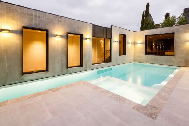 Modern minimalist courtyard exterior featuring a rectangular swimming pool surrounded by pale stone paving. The concrete walls have dark-framed windows and doors with warm interior lighting, plus a recessed illuminated opening resembling an outdoor bar or service window. Tall narrow trees rise behind the walls, and integrated wall lighting creates a warm evening atmosphere.