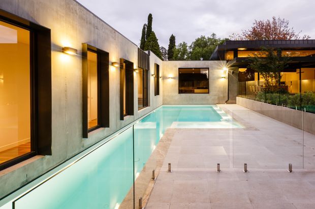 A contemporary courtyard residence viewed from the pool terrace at dusk. A long concrete wall with black-framed windows and warm architectural lighting borders a narrow turquoise swimming pool, while a pale stone-paved terrace leads toward a glazed pavilion and landscaped courtyard. The composition highlights clean geometric forms, indoor-outdoor connection, and concealed evening illumination.
