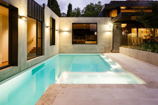A contemporary residential courtyard at dusk featuring a rectangular swimming pool surrounded by pale stone paving. The pool is enclosed by smooth rendered walls with large dark-framed windows and warm interior lighting, while a raised terrace on the right contains an outdoor dining area beneath deep roof overhangs.