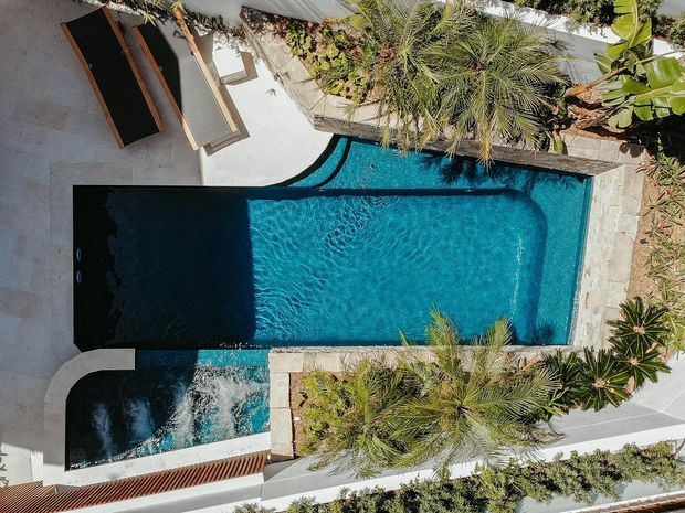 Overhead view of a contemporary landscaped pool area featuring a long turquoise swimming pool, an attached dark plunge pool or spa with a small connecting spillway, pale stone paving, timber sun loungers, and lush tropical planting enclosed by light rendered walls.
