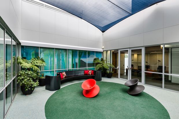 A bright contemporary indoor courtyard or atrium lounge with white wall cladding, extensive glazed partitions, abundant potted greenery, a curved black built-in sofa, and sculptural red and black lounge chairs. A dramatic blue translucent canopy forms the ceiling, while green carpet defines the central seating area.