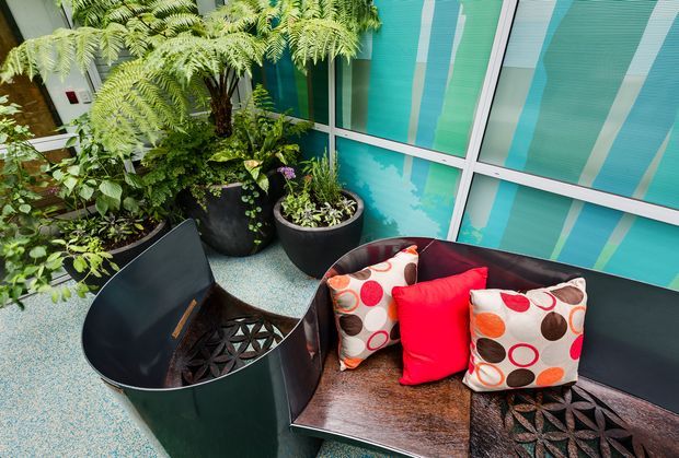 A compact indoor garden lounge or courtyard-like seating area with lush potted ferns and leafy plants, a sculptural dark metal lounge chair, and a built-in dark bench with colourful patterned cushions. Turquoise patterned glazing forms the backdrop, creating a vibrant contemporary interior with strong botanical and geometric elements.