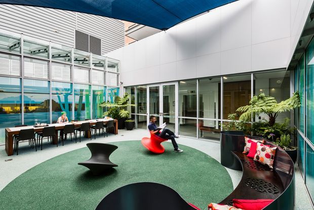 A contemporary commercial courtyard or atrium lounge beneath a blue tensile canopy. The space features white panelled walls, extensive glazed façades and doors, a green carpeted seating area, modern black sculptural furniture, a bright red lounge chair, curved dark benches with colourful cushions, potted plants, and an outdoor dining area along the glazed wall.