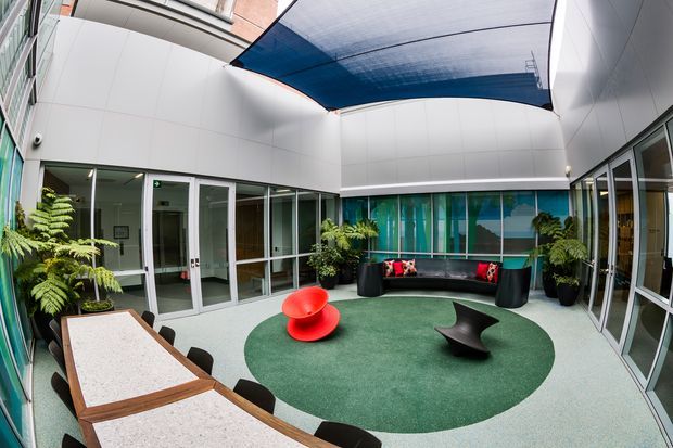 A contemporary enclosed courtyard or atrium with white wall panels, extensive glazed doors and windows, a curved translucent blue overhead canopy, and indoor planting. The space includes a green circular carpet, black built-in style seating, a bright red lounge chair, a dark sculptural coffee table, and a stone-topped table with chairs. It appears to be a polished commercial or institutional interior courtyard designed as a social breakout area.