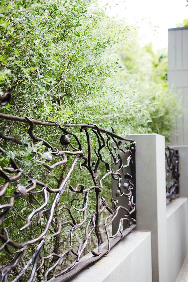 Close-up exterior view of a contemporary boundary or balcony railing with an artistic, irregular wrought-metal design, mounted on pale concrete or rendered masonry walls. Dense leafy vegetation fills the background, creating a garden-like setting. The image is portrait-oriented and focuses on the decorative balustrade and exterior architectural details.