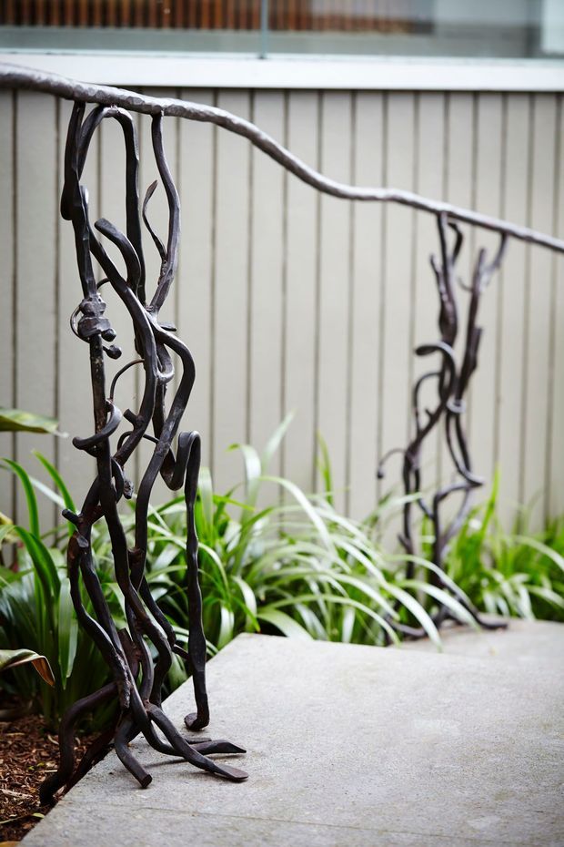 Close-up view of an organic, branch-like dark metal garden railing beside a pale concrete walkway. Lush strappy planting sits against a light vertical-panel boundary wall, creating a naturalistic outdoor courtyard or garden setting.