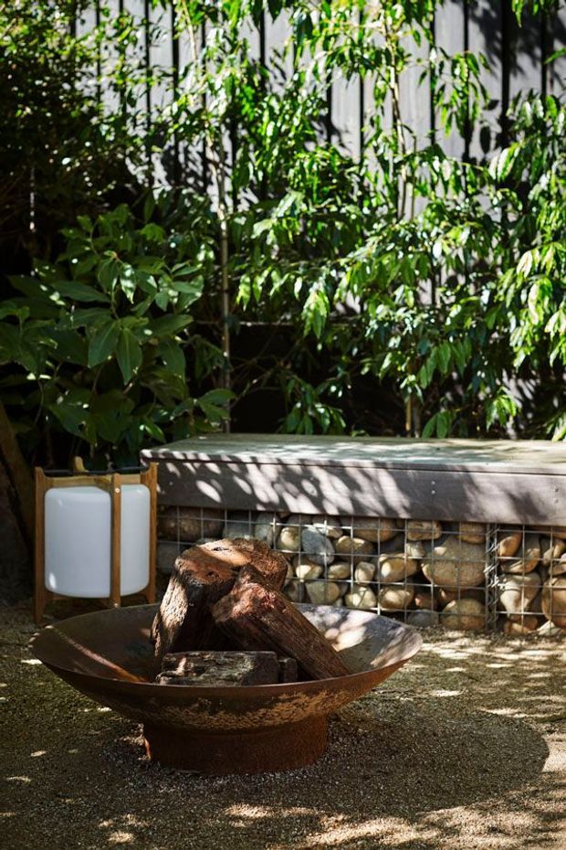 A shaded outdoor courtyard or garden area featuring a rusted metal fire bowl with logs, positioned in front of a gabion-style stone retaining wall. Dense tropical greenery surrounds the space, with dappled sunlight filtering through the foliage and a small outdoor lamp beside the fire bowl.