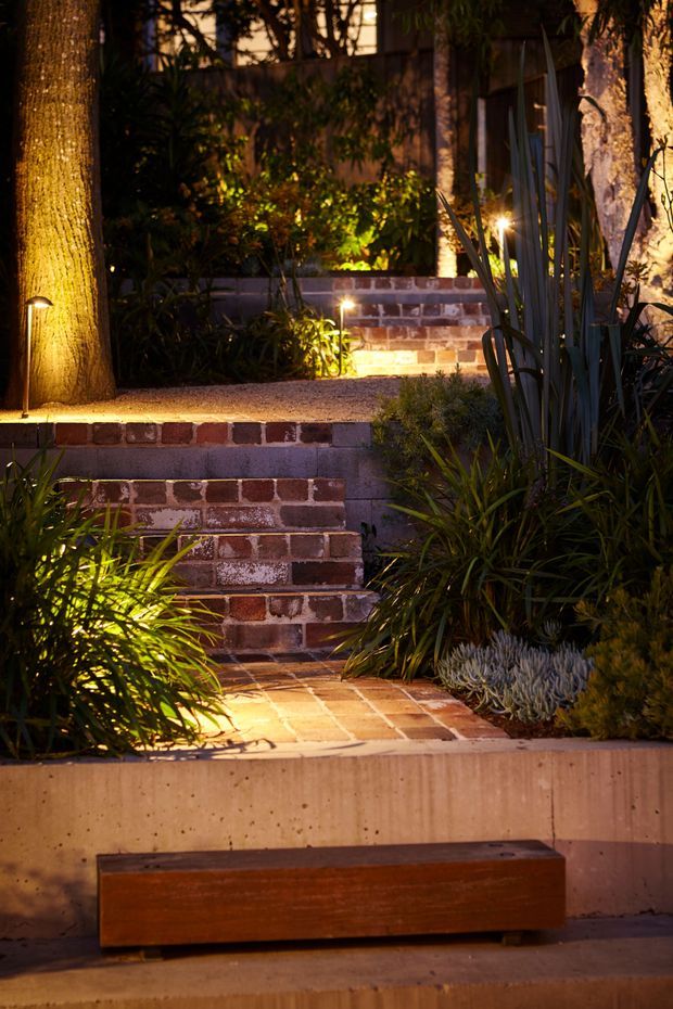 A warmly lit landscaped garden entrance at dusk, featuring brick retaining walls and steps, a brick-paved path, concrete garden edging, dense ornamental planting, mature trees, and subtle integrated landscape lighting. A simple timber bench sits in the foreground.