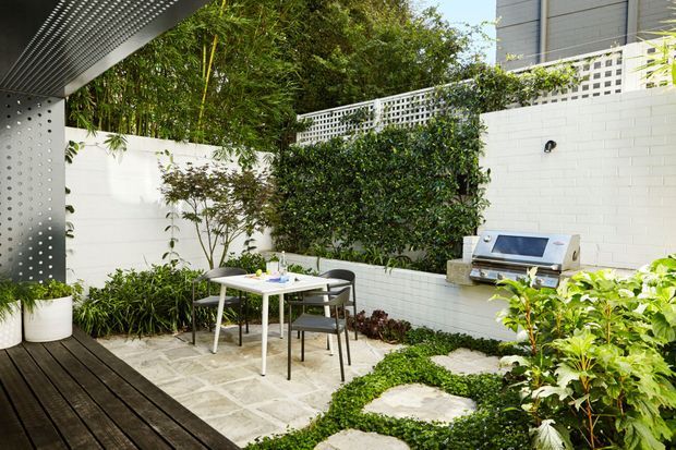 A compact landscaped courtyard or patio beside a contemporary building, featuring a timber deck, pale stone paving, white masonry boundary walls, dense green planting, an outdoor dining table with chairs, and a built-in outdoor barbecue. A perforated metal canopy or screen frames the upper left corner.
