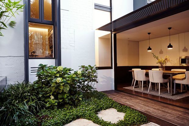A contemporary courtyard viewed from a planted garden, with white masonry walls, dark-framed windows, timber decking and a covered outdoor dining area. The dining space contains a long table, upholstered chairs, pendant lights and a kitchen visible at the rear. Lush greenery softens the compact courtyard setting.