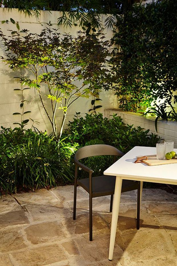 A small landscaped courtyard or patio with dense planting against pale rendered and tiled walls. A white outdoor dining table with a dark chair sits on irregular natural stone paving, creating an intimate, leafy dining setting.