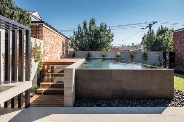 Contemporary urban backyard courtyard with a raised rectangular swimming pool finished in dark stone or concrete, integrated timber steps and decking, rendered boundary walls, exposed brick walls, and vertical metal screening. Mature planting and neighbouring rooftops surround the bright outdoor space.