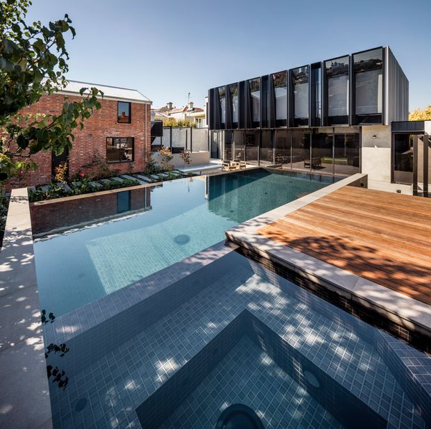 A contemporary urban residential courtyard featuring a large tiled swimming pool with integrated stepped sections, a timber deck, and a modern two-storey extension. The extension has dark vertical cladding, extensive glazing, and a rectilinear form, contrasting with the adjoining red-brick heritage-style dwelling. Mature planting frames the pool area.