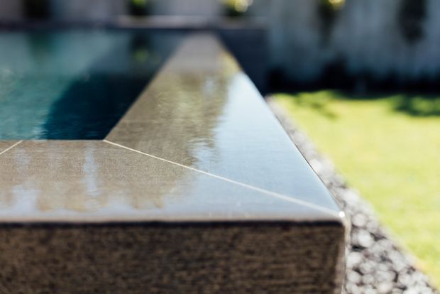 Close-up detail of a contemporary swimming pool edge with a dark stone or tiled coping surface, reflective water to the left, and a gravel border with lawn beyond. The shallow depth of field emphasizes the crisp pool-edge construction and material contrast.