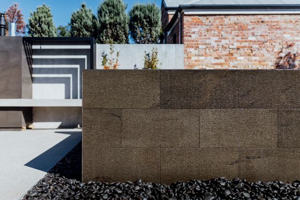A contemporary exterior courtyard or garden composition featuring a dark textured masonry privacy wall, a geometric dark facade element with layered white horizontal bands, pale paving, black decorative stone, and an exposed red-brick building wall with a dark metal roof edge. Mature trees rise behind the walls.