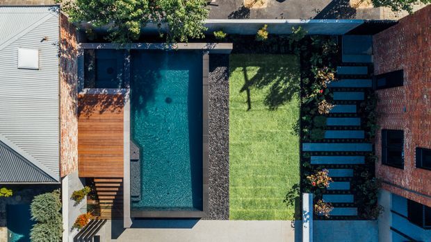 Aerial view of a compact residential courtyard showing a rectangular swimming pool, timber pool deck, manicured lawn, dark stepping-stone path, planted borders, and surrounding brick and metal-roofed building volumes.