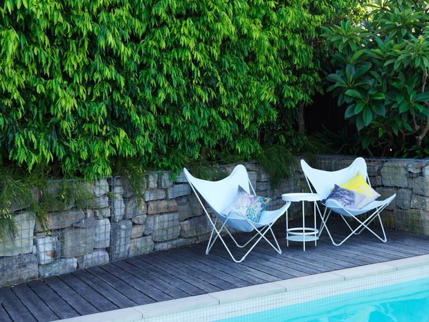 A leafy outdoor poolside terrace with dark timber decking, a turquoise swimming pool, a low gabion-style stone retaining wall, and two white sling lounge chairs with cushions around a small white side table. Dense greenery creates a private garden setting.