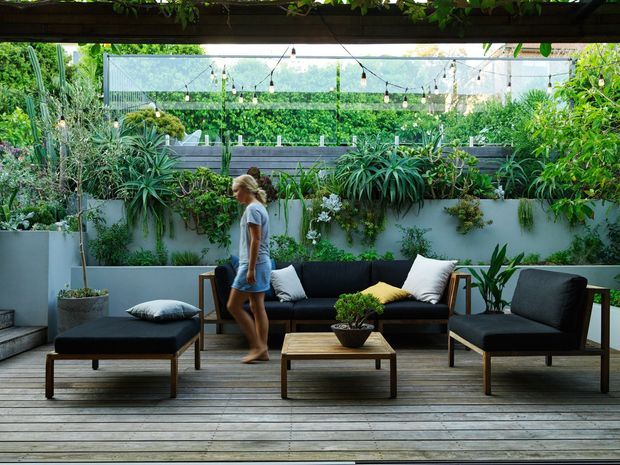 A lush, covered outdoor lounge on a timber deck, furnished with a black sofa, matching armchairs, ottoman, and low coffee table. Dense vertical and container planting surrounds the space, with string lights suspended beneath the overhead timber structure.
