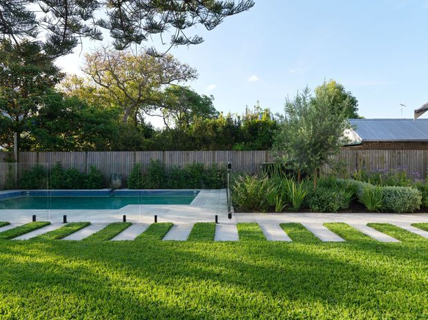 A landscaped residential backyard featuring a rectangular swimming pool, pale paved stepping paths through a broad lawn, dense planting beds, and a timber privacy fence. A low building with a corrugated metal roof is partially visible behind the garden, with mature trees and clear blue sky beyond.