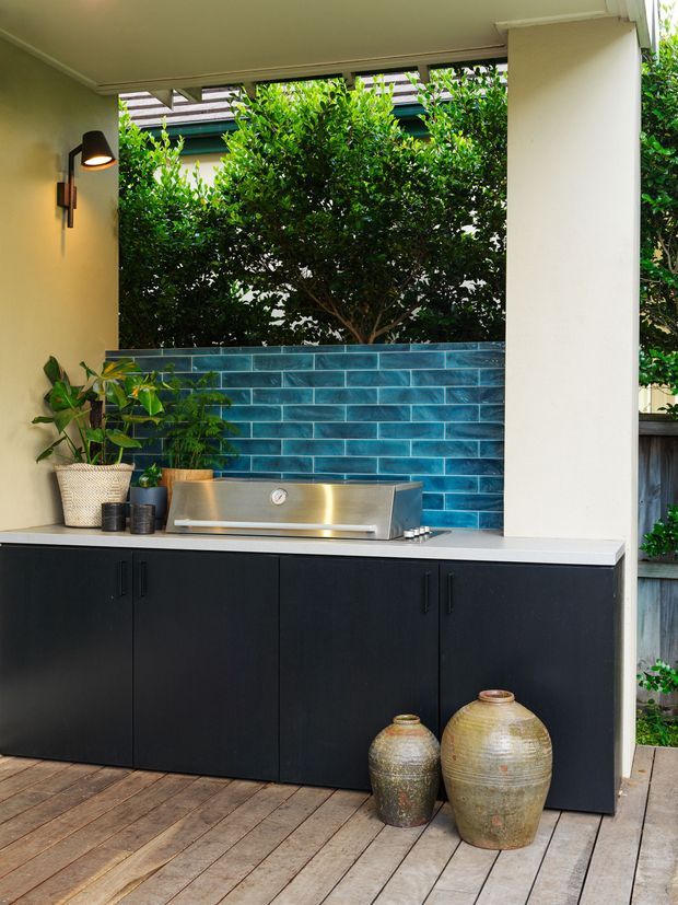 A compact covered outdoor kitchen on a timber deck, featuring a stainless-steel built-in barbecue set into matte black cabinetry, a pale benchtop, and a vivid blue glazed-tile splashback. Dense greenery is visible through the open wall, with an exterior wall light, potted plants, and decorative ceramic vessels adding detail.