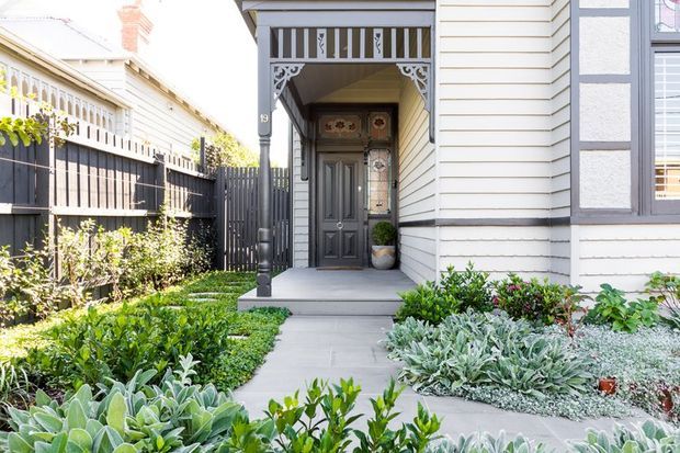A bright view of a traditional home entryway with a covered porch, decorative timber fretwork, a dark paneled front door with stained-glass inserts, white horizontal weatherboard cladding, and a concrete path through a densely planted front garden. A dark timber boundary fence and neighboring porch are visible to the side.