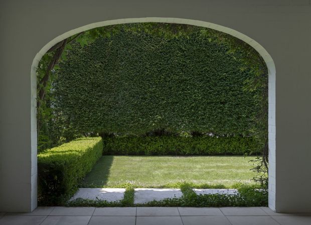 A symmetrical view from a tiled interior or covered passage through a broad arched opening, framing a manicured garden. The opening has pale rendered walls and a light stone or painted masonry arch, with clipped hedges, a dense green hedge backdrop, lawn, and a simple stepping-stone path. The composition is calm and strongly focused on the architectural garden connection.