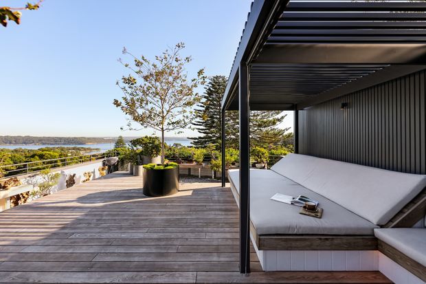 A spacious elevated outdoor terrace with timber decking, a large built-in upholstered daybed beneath a dark slatted pergola, and vertical dark wall cladding. Potted trees and coastal water views are visible beyond the terrace railing, creating a refined outdoor entertaining and relaxation space.
