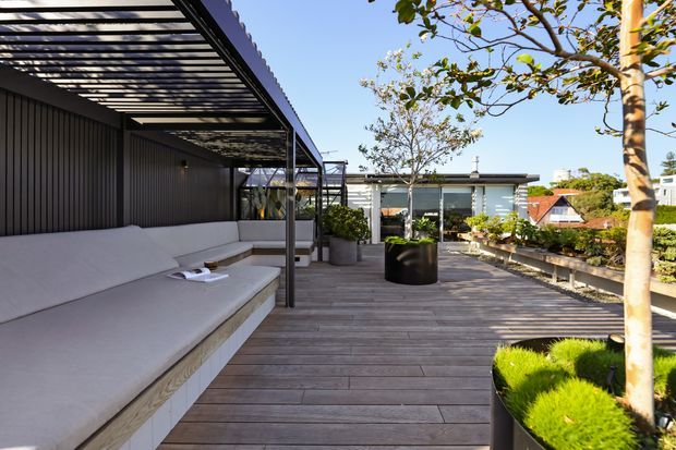 A contemporary rooftop-style outdoor terrace with a broad timber deck, built-in upholstered bench seating beneath a black metal pergola, vertical dark wall cladding, large planters, and landscaped greenery. The terrace overlooks neighbouring rooftops and includes glazed architectural elements in the background.