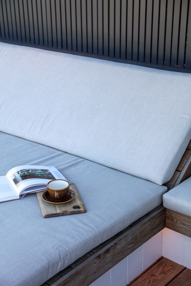 Close-up of a built-in outdoor lounge daybed or bench with pale blue-grey upholstered cushions, a dark vertical slatted backrest, and weathered timber framing. An open magazine and coffee cup sit on a wooden tray, creating a relaxed outdoor seating scene.