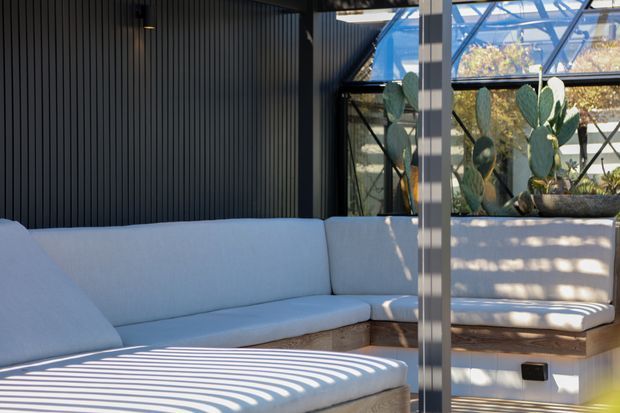 A bright, contemporary sunroom or enclosed outdoor lounge with a built-in pale upholstered sectional bench, warm timber base, dark vertical wall cladding, and a glazed roof and wall structure. A large cactus planter sits behind the seating, while strong striped shadows from the glazing create a graphic pattern across the cushions.