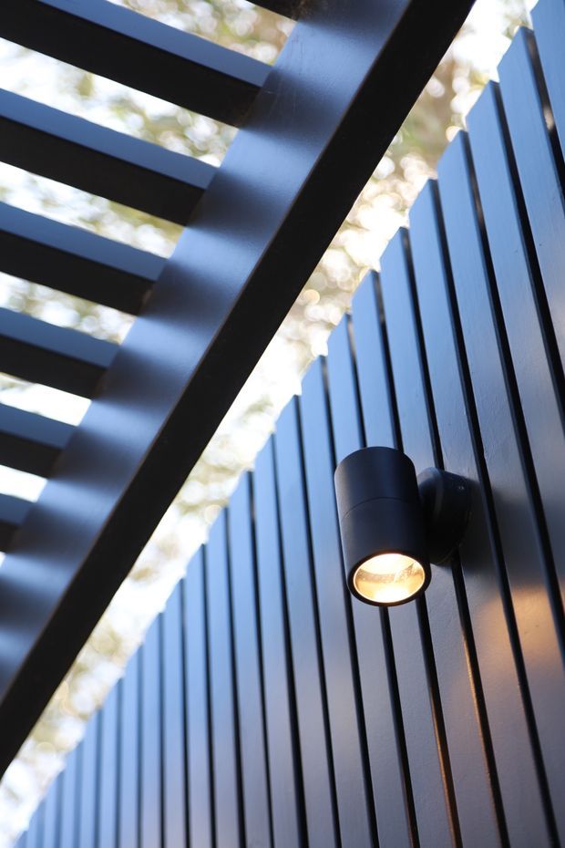 Close-up architectural detail of a dark exterior wall clad in narrow vertical boards, featuring a cylindrical wall-mounted light and a heavy black slatted overhead structure. Bright foliage is softly visible beyond the open slats, creating a high-contrast, contemporary outdoor facade composition.