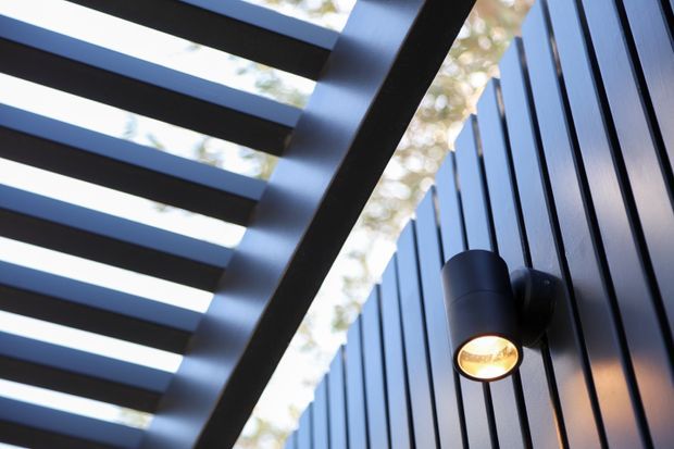 Close-up architectural detail of a dark exterior wall with vertical ribbed cladding and a black cylindrical wall-mounted light. A slatted overhead structure creates strong linear shadows, with blurred trees visible beyond.