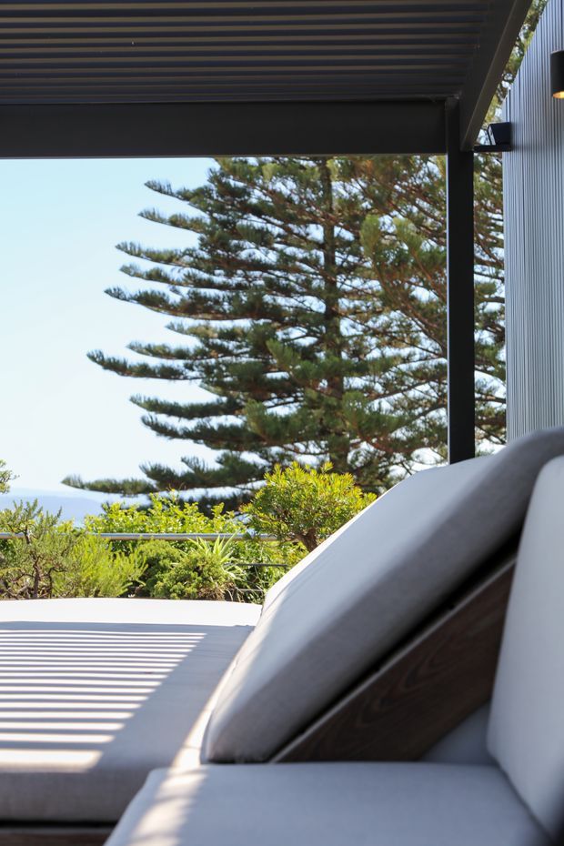 A sunlit outdoor terrace or balcony with a built-in upholstered lounge seat beneath a dark louvred pergola. The space features dark structural framing, a ribbed grey exterior wall, timber detailing on the seating, and landscaped planting with a large Norfolk Island pine beyond.