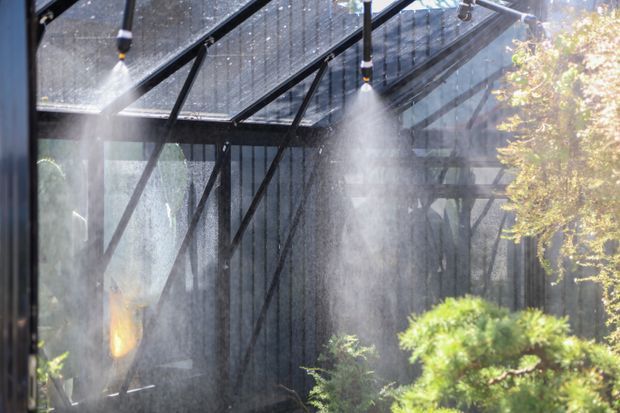 A glass-enclosed garden structure with a dark metal frame and transparent pitched roof, fitted with overhead misting or irrigation nozzles. Fine water spray fills the space around lush green plants, creating a humid conservatory or greenhouse atmosphere.
