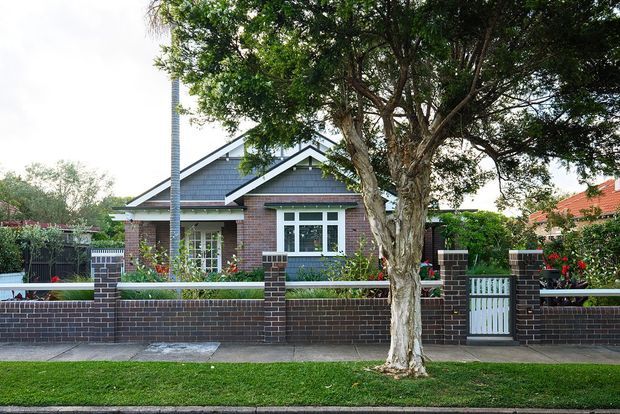 Street-facing view of a single-storey brick bungalow with a dark pitched gable roof, white trim, front porch, landscaped garden, and a low brick-and-metal boundary fence. A mature tree partially screens the facade.