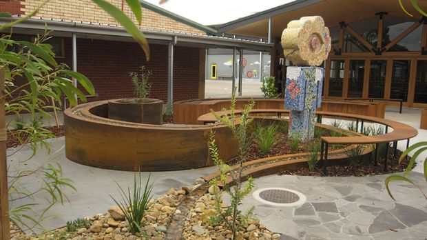 A landscaped interior courtyard or atrium with curved rust-toned steel planter walls, built-in timber seating, native-style planting, stone paving, and a colourful sculptural column. Glazed doors and brick walls surround the sheltered communal space.