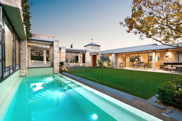 A contemporary courtyard-style residence viewed from beside a long turquoise swimming pool. The low, flat-roofed building is arranged around a manicured central lawn, with extensive glazing, pale stone masonry, timber-framed openings, covered outdoor living areas, and a small pavilion-like roof element. Glass pool fencing separates the pool from the lawn, while mature planting and autumn foliage frame the scene.