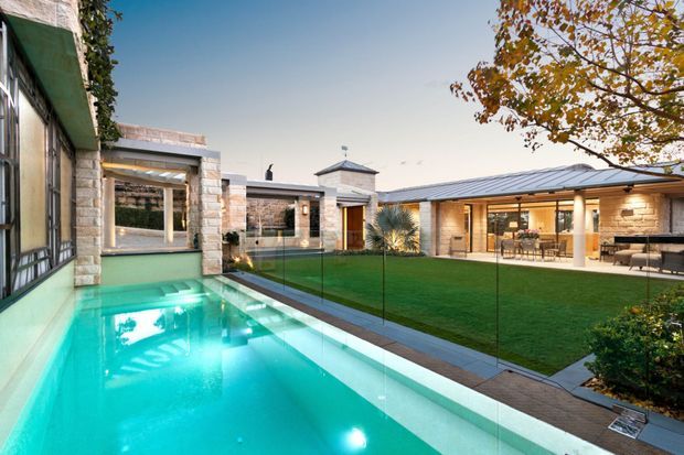 Contemporary single-level courtyard residence arranged around a narrow illuminated swimming pool and manicured lawn. The home features pale stone masonry, broad glazed openings, slim dark metal frames, a covered outdoor entertaining area, and a small pavilion-like roof element. Clear frameless pool fencing separates the pool from the central courtyard, with mature planting and autumn foliage around the perimeter.