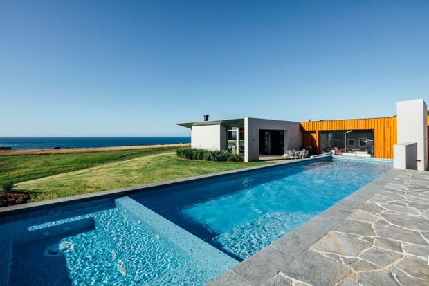 A contemporary single-storey coastal home viewed from its swimming pool. The low, rectilinear building has white rendered volumes, dark-framed glazing, and a prominent orange timber-clad section. A large blue pool and shallow spa occupy the foreground, bordered by irregular grey stone paving, with a grassy lawn and ocean beyond.