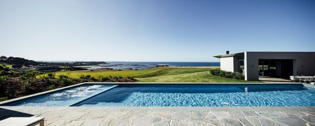 Wide exterior view of a minimalist contemporary single-storey home beside a swimming pool, with a flat-roofed dark and white facade, expansive glazing, pale stone pool terrace, and an open coastal landscape with ocean beyond.