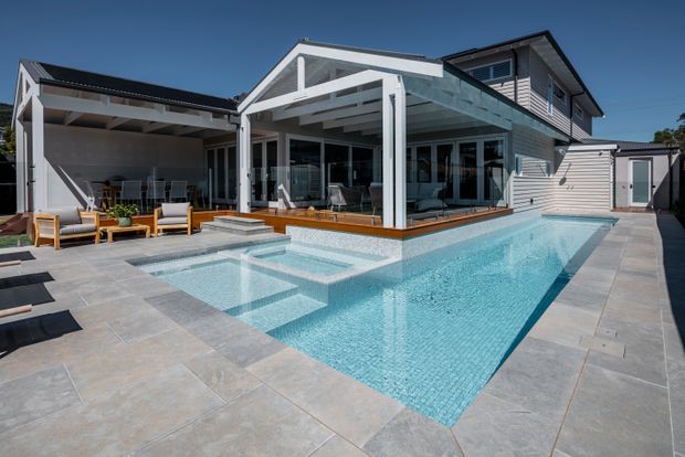 A contemporary multi-level home viewed from the rear, with a large rectangular swimming pool, broad paved terrace, covered outdoor entertaining area, and extensive glazed living spaces. The facade combines light horizontal cladding, dark roofing, and a prominent gabled covered structure.