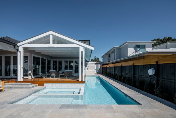 A bright contemporary residential outdoor area featuring a long narrow swimming pool alongside a covered patio and glazed pavilion. The pool terrace uses pale stone-like paving, with timber decking, white structural framing, extensive glass doors, and a dark vertical fence with a timber privacy screen. Modern neighboring homes and a clear blue sky are visible beyond.