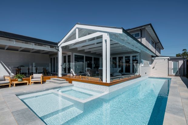 Bright exterior view of a contemporary coastal-style home centered on a covered glass outdoor living pavilion overlooking a tiled swimming pool. The house has white weatherboard-style cladding, dark pitched roofs, extensive glazing, timber decking, paved pool surrounds, and a connected outdoor dining/lounge area.