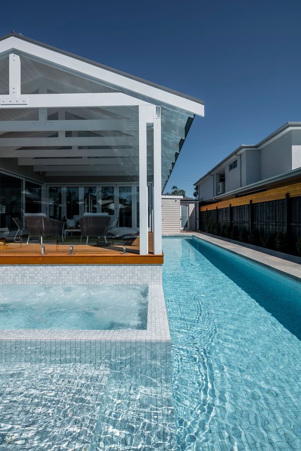 Bright exterior architectural photograph of a contemporary white gabled pavilion opening onto a timber deck and a long narrow swimming pool. The pool includes a raised spa or shallow water feature in the foreground and pale blue mosaic tiling throughout. Glazed doors, exposed white structural rafters, and a covered outdoor seating area face the pool, with neighbouring modern homes and a dark timber fence visible alongside.