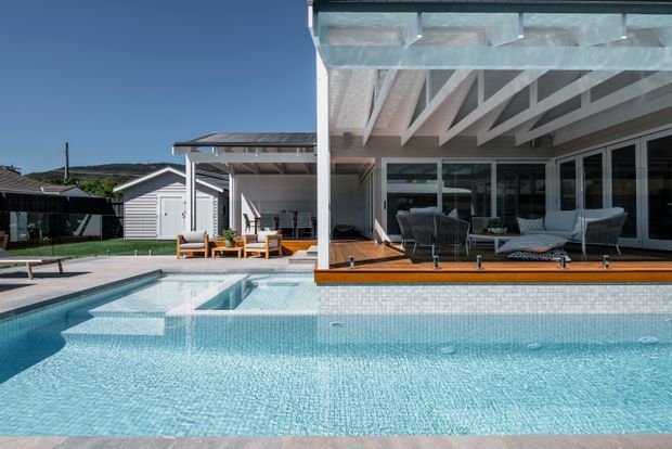 Contemporary residential pool area beside a glazed outdoor entertaining pavilion. The image shows a large pale blue tiled swimming pool, timber decking, white-framed sliding glass doors, a covered terrace with exposed white-painted rafters, and outdoor lounge furniture. A small lawn, detached light-coloured outbuilding, and neighbouring suburban structures are visible in the background.