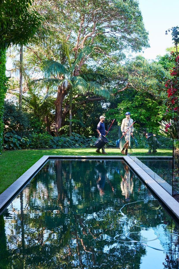 A long rectangular swimming pool set within a lush, densely planted garden. The pool has dark reflective water and crisp pale paving edges, with tropical foliage and mature trees surrounding it. Two people are visible in the background tending the garden, while reflections of the vegetation fill the pool surface.