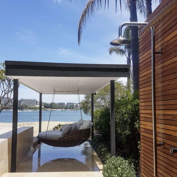 A modern waterfront outdoor shower and covered lounge area beside a timber-clad wall. The space features a suspended woven hanging chair, dark slender posts, a flat roof with a white-lined soffit, reflective stone paving, dense planting, and a stainless steel rain shower. A sandy shoreline, water, and buildings are visible beyond.