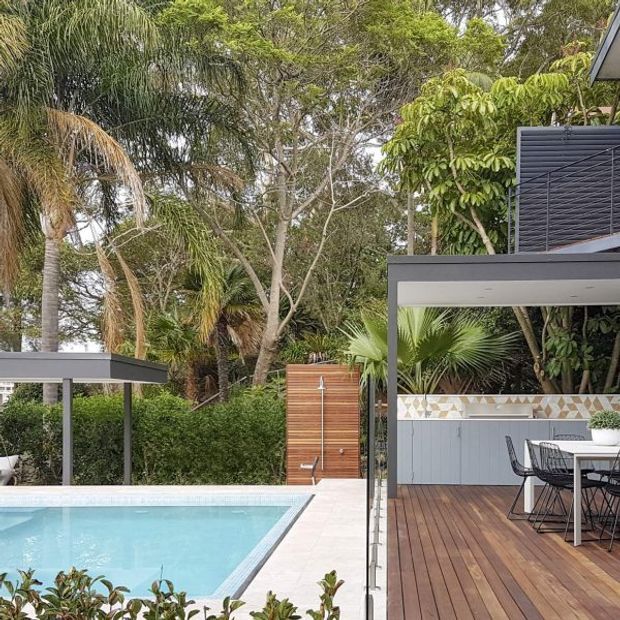 A lush tropical outdoor entertaining area with a rectangular swimming pool beside a timber deck. The deck includes a dining table and chairs, a built-in outdoor kitchen or counter with a geometric tiled splashback, and a vertical timber outdoor shower. Modern dark-framed structures and screening contrast with dense palms and landscaped greenery.