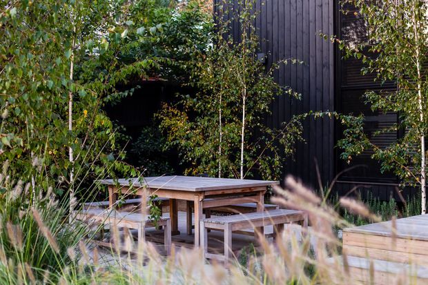 A landscaped outdoor dining area with a timber picnic-style table and bench seating, surrounded by dense planting and ornamental grasses. A dark vertical timber-clad building forms the backdrop, with a raised timber deck or platform visible at right.