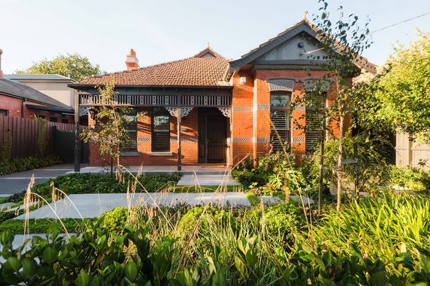 Street-facing view of a heritage red-brick bungalow with a terracotta tiled roof, decorative timber veranda, ornate gable detailing, and leadlight-style windows. The home is set behind a lush, informal front garden with a paved walkway.